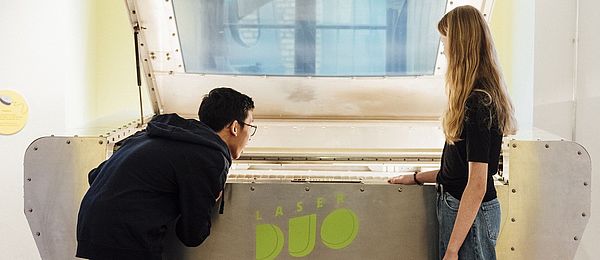 Ein Mädchen und ein Junge betrachten ein großes, silberfarbenes technisches Gerät. Auf dem gerät steht „LaserDuo“. Der Junge beugte sich über die Öffnung des Geräts, das Mädchen steht neben ihm und betrachtet das Innere des Geräts.