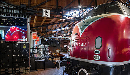 Die Schnellzuglokomotive V 200 steht in den historischen Lokschuppen des Deutschen Technikmuseums auf in den Holzboden eingelassenen Schienen. Ihr abgerundeter Rumpf ist Feuerrot lackiert. Links daneben befindet sich ein Neonschild in Form der Lokomotive.