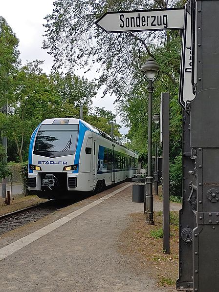 Ein blau-weißer Zug mit der Aufschrift "Stadler" fährt auf einem Bahnsteig im Grünen ein, rechts ein Schild mit der Aufschrift "Sonderzug".