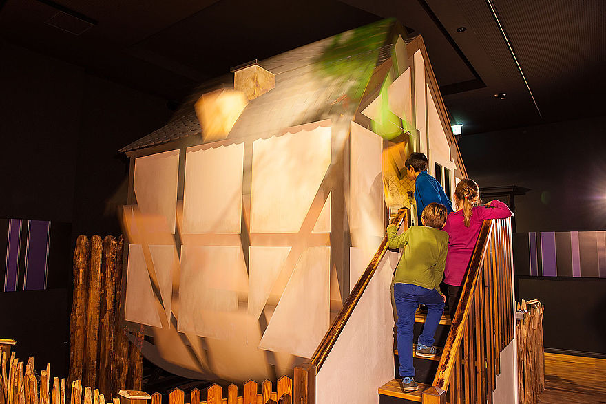 In einem Raum befindet sich hinter einem Holzzaun ein kleines Haus. Drei Kinder betreten das Haus über eine Treppe.