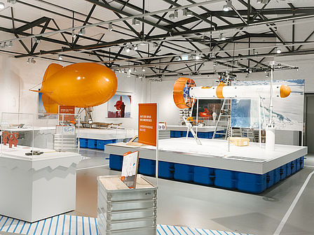 Blick in die Ausstellung: Links ein orangener Fesselballon, rechts eine lange Messsonde. Die Ausstellungsmodule liegen auf "Eisschollen".