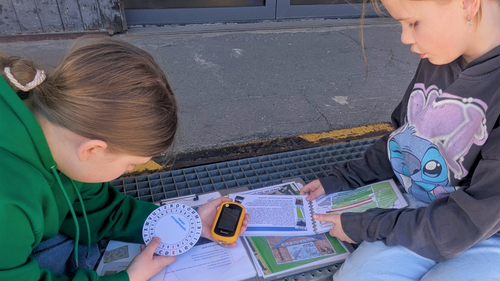 Zwei Kinder sind über die Materialen zur GPS-Tour gebeugt. Ein Kind hält eine Verschlüsselungsscheibe in der Hand.