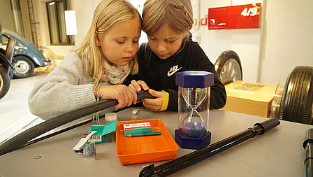 Ein Mädchen und ein Junge im Grundschulalter suchen gemeinsam nach einem Loch im Fahrradschlauch. Vor ihnen stehen ein Kästchen mit Flickzeug und eine Sanduhr. 