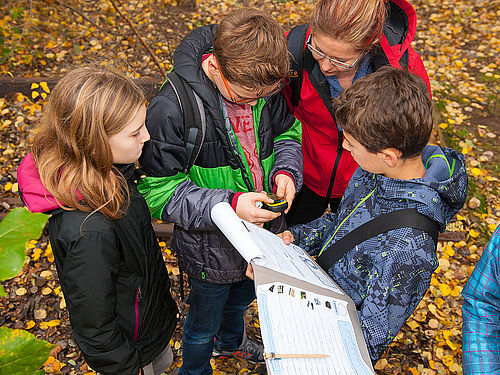 Vier Kinder und eine Betreuerin stehen auf einem belaubten Untergrund. Eines der Kinder hält eine geöffnete Klemmmappe mit Quiz-Fragen in der Hand. Ein anderes Kind gibt Koordinaten in ein kleines GPS-Gerät ein.