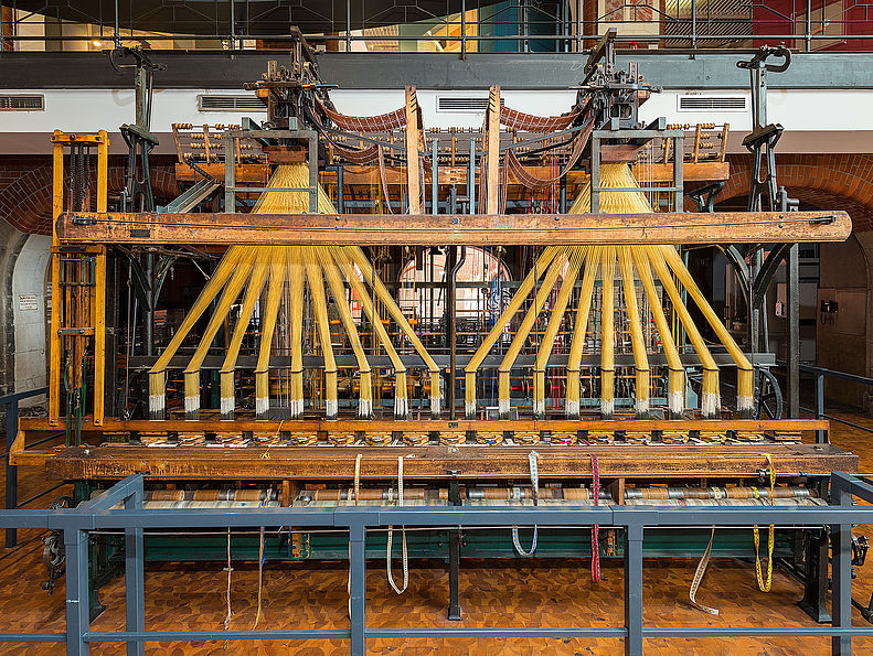 Der Webstuhl ist etwa fünf Meter breit, 2,4 Meter tief und 3,5 Meter hoch. Die beiden Webstuhlaufsätze sind oben an einem Metallrahmen aufgehängt. Sie sehen aus wie zwei riesige Rechen mit je acht Zähnen.