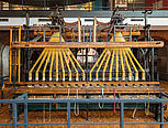 Der Webstuhl ist etwa fünf Meter breit, 2,4 Meter tief und 3,5 Meter hoch. Die beiden Webstuhlaufsätze sind oben an einem Metallrahmen aufgehängt. Sie sehen aus wie zwei riesige Rechen mit je acht Zähnen.
