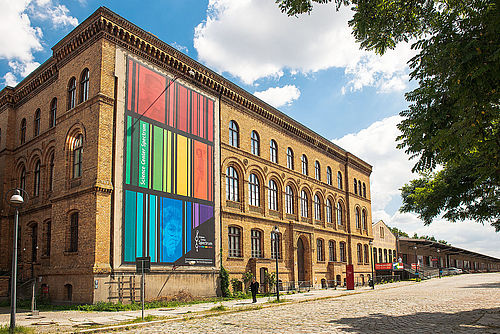 Ein dreigeschossiger Backsteinbau mit einem bunt gestreiften Transparent an der Fassade. Davor eine kopfsteingepflasterte Straße.