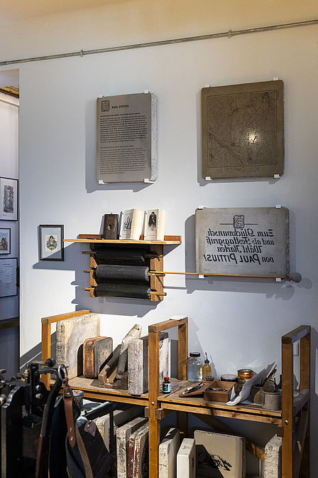 Das Foto zeigt eine Wand, an der mehrere Lithografiesteine hängen, einer davon ist eine Gedenktafel für die Brüder Gerson, Darunter sieht man ein Regal mit verschiedenen Werkzeugen für den Steindruck.