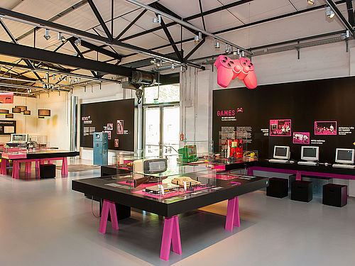Die Themeninsel Games in der Ausstellung „Das Netz“. Sie besteht aus schwarzen Tischen mit pinken Tischbeinen. Auf den Tischen befinden sich Glasvitrinen mit Objekten. Über den Tischen hängt einer großer, pinkfarbener Gamecontroller.