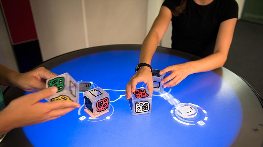 An einem Tisch mit einer runden Oberfläche befinden sich zwei Personen. Die Oberfläche hat außen einen dunklen Ring und innen leuchtet sie blau. Auf der blauen Fläche liegen eine Scheibe und drei etwa faustgroße Würfel mit farbigen Mustern. Die linke Person hält einen weiteren Würfel in den Händen. Die rechte Person greift zwei der Würfel auf dem Tisch. Unter den Würfeln und der Scheibe und direkt um sie herum leuchten weiße Muster.