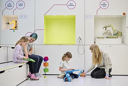 Mehrere Kinder und Erwachsene sitzen auf den Sitzstufen und auf dem Boden eines hell gestrichenen Raums. Sie lesen Bücher und beschäftigen sich mit kleinen Robotern.