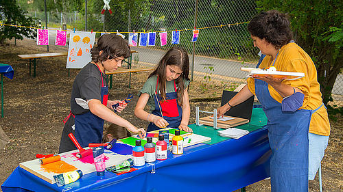 Zwei Kinder und eine Frau stehen an einem Tisch und drucken mit bunten Farben Pflanzenmotive. Im Hintergrund hängen fertige Drucke an einer Leine zum Trocknen.