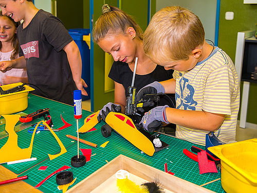Vier Kinder kleben an einem Tisch kleine Autos aus Pappe mit einer Heißklebepistole zusammen. Auf dem Tisch liegen grüne Arbeitsmatten, bunte Pappschnipsel, gelbe Schalen und Klebestifte. 