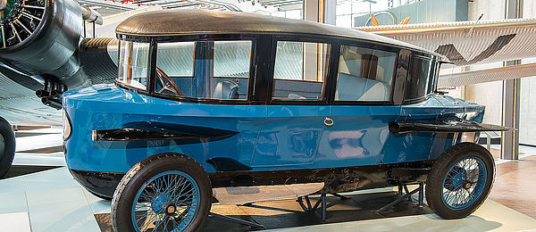 Ein historisches, längliches, blaues Fahrzeug, das durch seine aerodynamische, tropfenähnliche Form auffällt. Das Dach fällt nach hinten ab, Scheinwerfer und Schutzbleche an den Seiten erinnern an Flugzeugflügel.