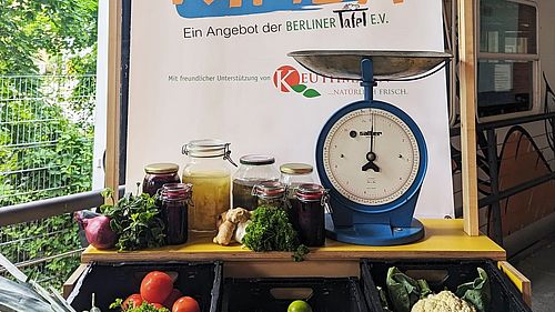 Vor dem KIMBA-Express steht eine Art Markstand mit einer blauen Gemüsewaage und verschiedenen Einweckgläseren mit eingelegtem Gemüse. 