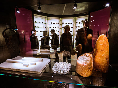 Fünf Personen stehen in einem Raum der Ausstellung „Alles Zucker!“. Im Vordergrund werden in einer Vitrine ein weiß-brauner Zuckerhut, Teile von Rohrzucker, Kandis-Zuckerkristalle und gepresster Zucker gezeigt. Im Hintergrund befinden sich deckenhohe Vitrinen mit Zuckerdosen.