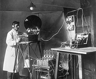 Ein Mann mit Kopfhörern bedient ein Gerät, das an ein Grammofon erinnert. Im Vordergrund sind viele Kabel und technische Geräte, die alle miteinander verbunden sind. 