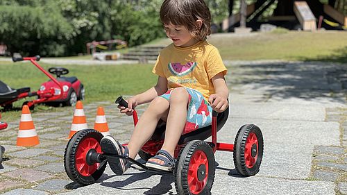 In the Museum Park a girl is riding a red three-wheel-vehicle.